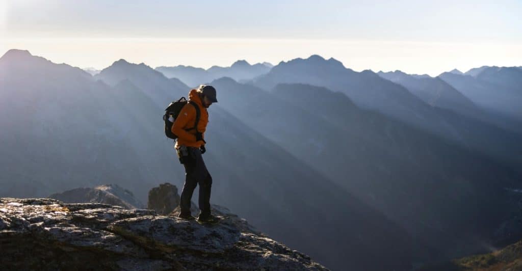  A person with a backpack is standing on a rocky mountaintop, looking out at a mountain range.