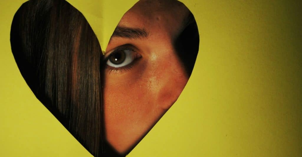 A close-up of a person's eye and part of their face visible through a heart-shaped cutout in yellow paper.