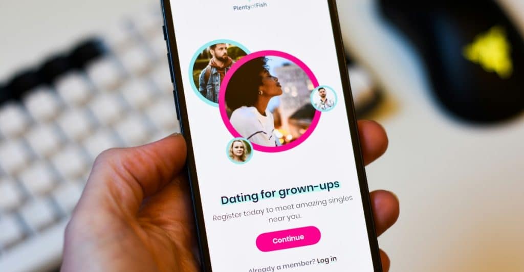 Close-up of a hand holding a smartphone displaying the Plenty of Fish dating app login screen with profile photos and text.