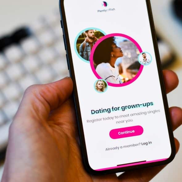 Close-up of a hand holding a smartphone displaying the Plenty of Fish dating app login screen with profile photos and text.