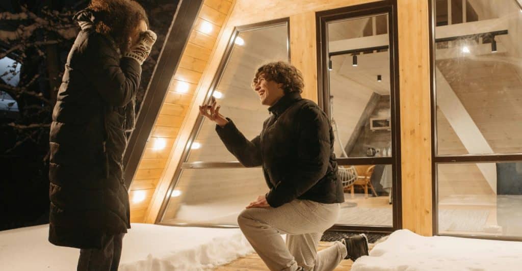 A man kneeling to propose to a woman in front of a snow-covered cabin at night.