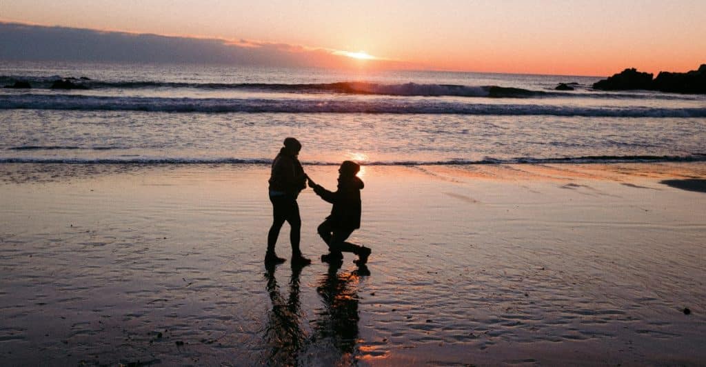A silhouette of a person kneeling to propose to another person on a beach at sunset.