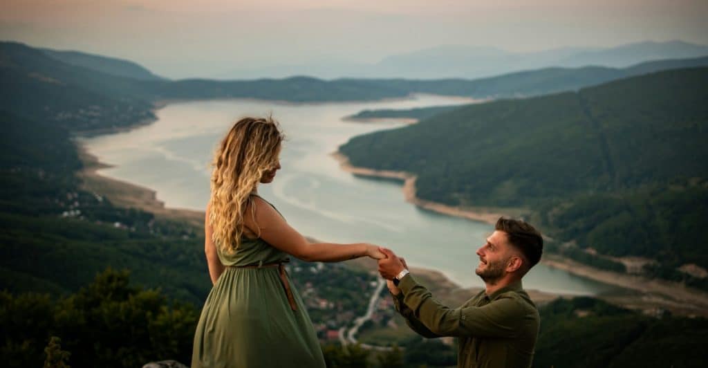  A man kneeling to propose to a woman on a mountain overlooking a lake at sunrise.