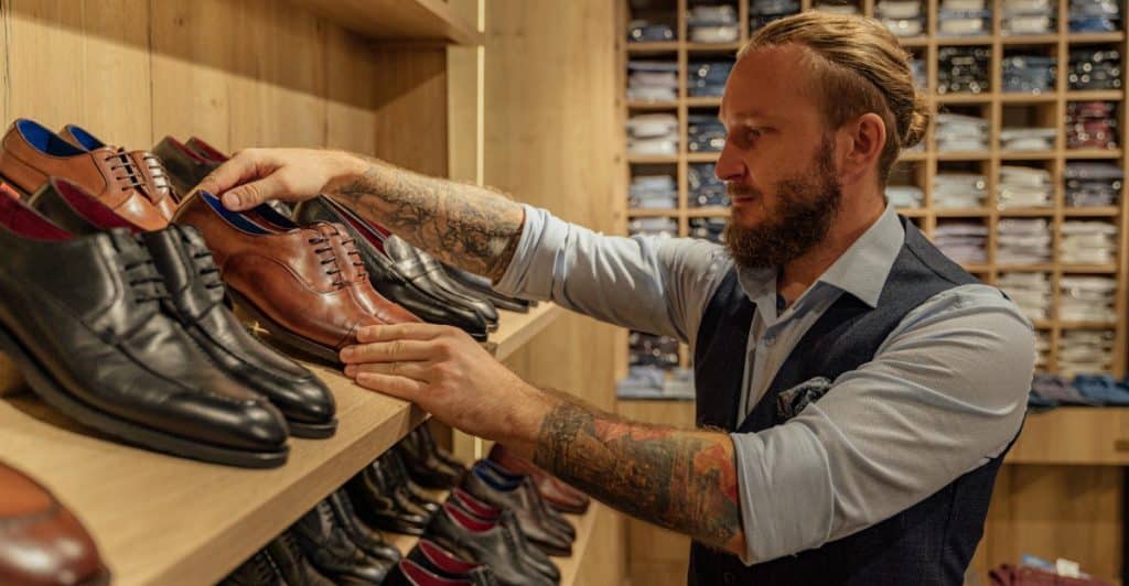 A man with arm tattoos, wearing a vest and light-colored shirt carefully looking at and selecting from a variety of men's dress shoes on a wooden shelf in a store.