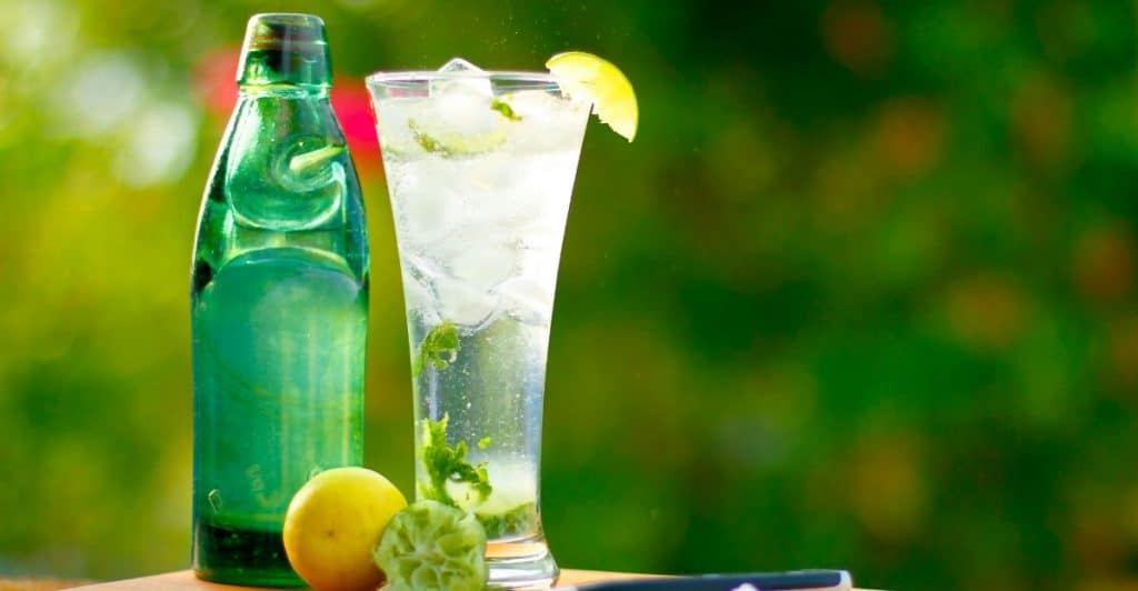 A tall glass of a clear, refreshing drink with ice, lime slices, and mint leaves sitting on a wooden cutting board next to a green bottle and a pile of salt.
