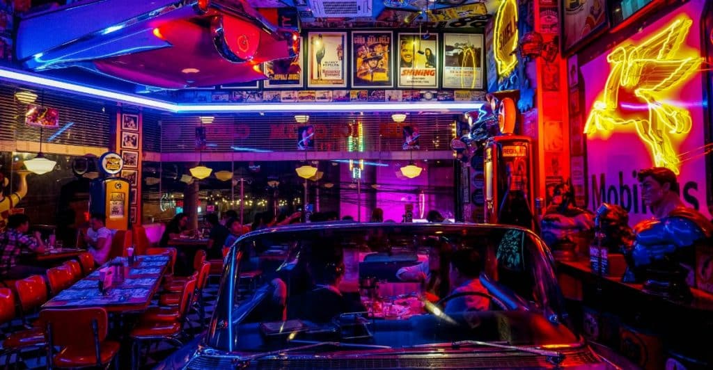 The interior of a retro-themed diner with neon lights, movie posters, booths, and part of a car.