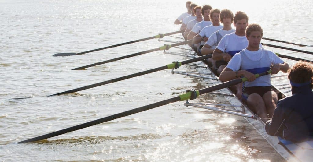 A rowing team of several men in white shirts rowing a boat on a body of water, with a person facing them in the stern.