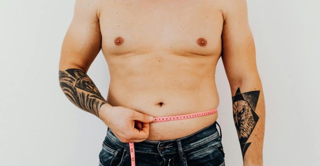 A shirtless man is measuring his waist with a pink measuring tape, with visible tattoos on his arms