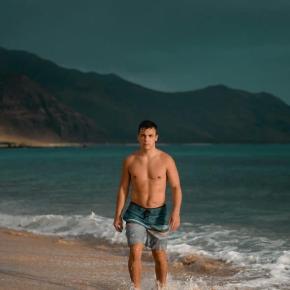 A shirtless man in swim trunks standing in water at the edge of a beach with waves crashing around his legs, with mountains in the background
