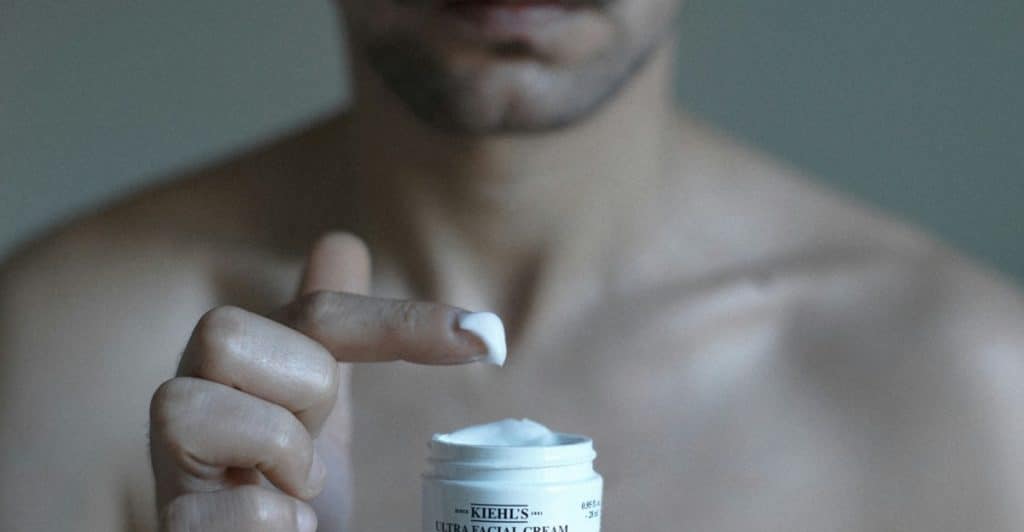 A close-up of a shirtless man's hands taking cream from a small white jar of Kiehl's facial cream.