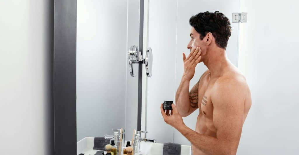 A shirtless man wearing a white towel around his waist applying a product to his face while looking in a mirror in a bathroom.