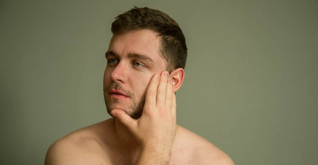 A close-up portrait of a shirtless man with brown hair and a light beard touching his face against a muted green background.