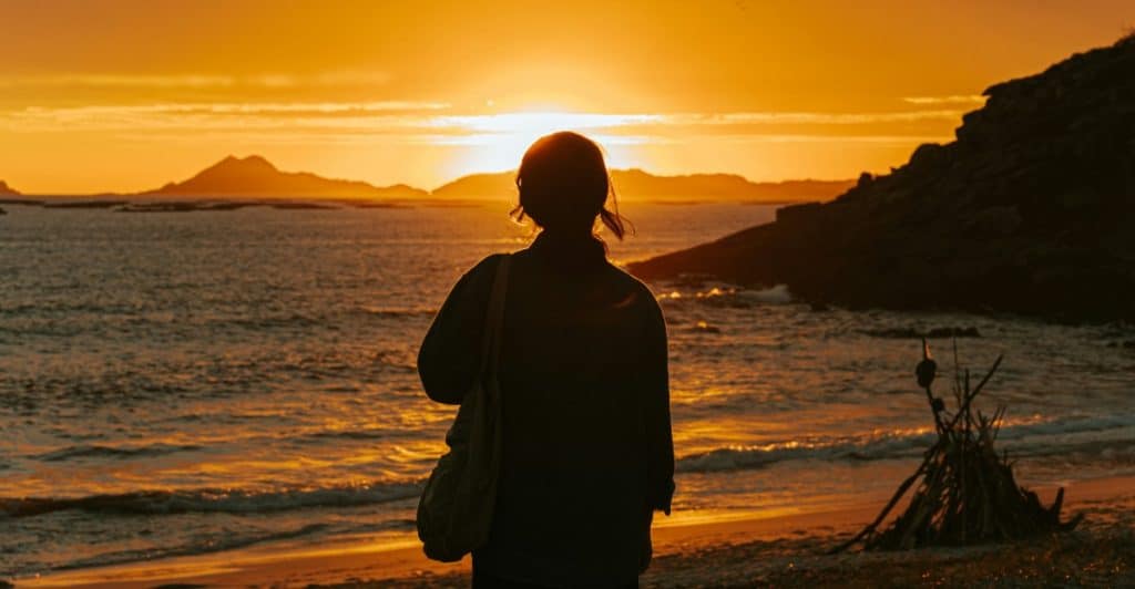 A person is silhouetted against a sunset over a body of water, with islands or land masses visible in the distance.