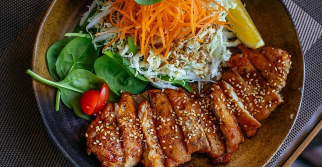 A plate of sliced chicken or meat with sesame seeds, served with a salad and a lemon wedge, illustrating a meal that includes greens.
