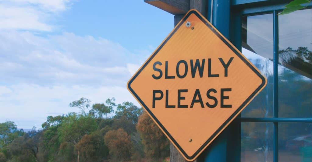 A yellow diamond-shaped road sign with the text "SLOWLY PLEASE" attached to a wooden post outdoors.