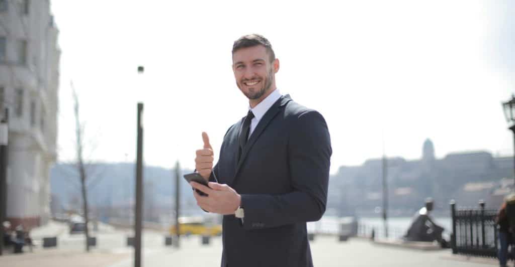 A smiling businessman in a suit and tie stands outdoors in a city, holding a smartphone and giving a thumbs-up gesture.