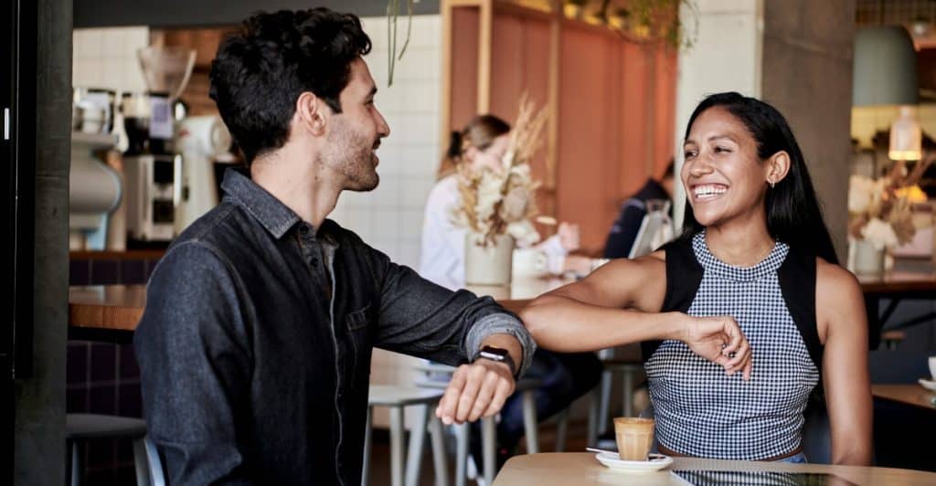 A man and a woman sit across from each other at a table in a cafe or bar, looking at each other and smiling.