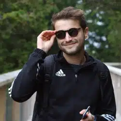 A man smiling while wearing sunglasses and a black jacket, holding a phone, standing outdoors on a wooden walkway with trees behind him.