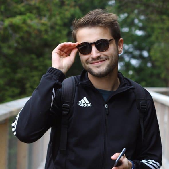 A man smiling while wearing sunglasses and a black jacket, holding a phone, standing outdoors on a wooden walkway with trees behind him.