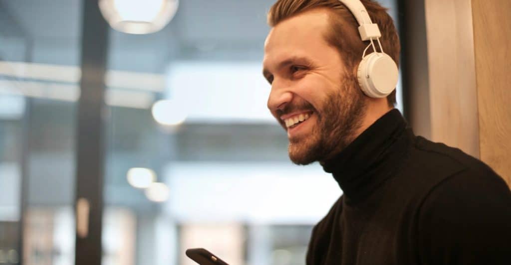 A smiling man with a beard and white headphones looking at his phone wearing a black turtleneck and standing in a modern indoor setting.