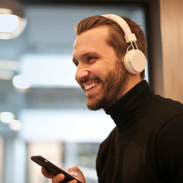 A smiling man with a beard and white headphones looking at his phone wearing a black turtleneck and standing in a modern indoor setting.