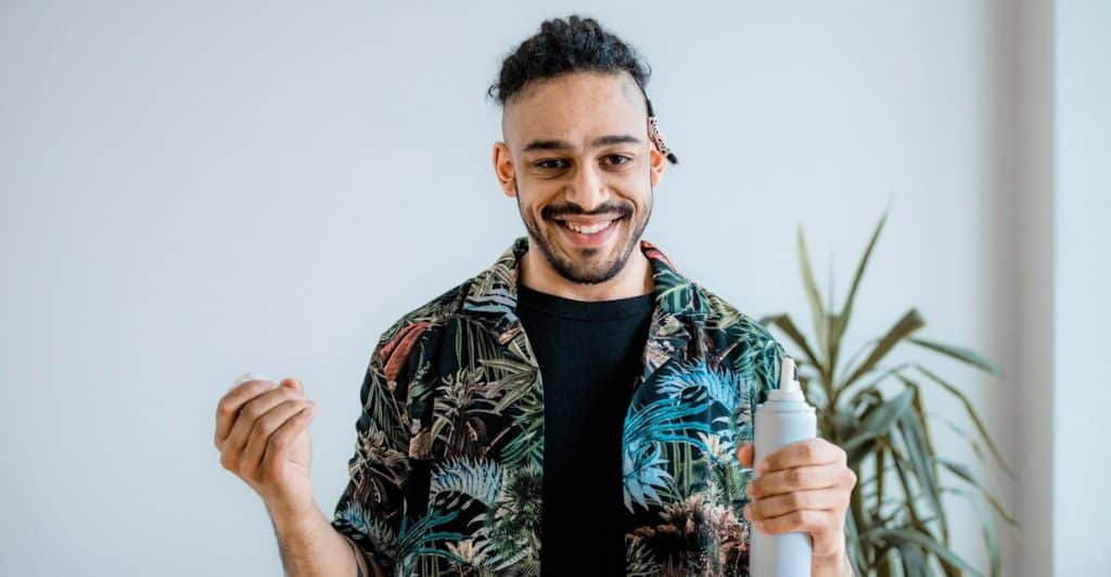 A man with dark curly hair, wearing a colorful shirt, holding a bottle of sunscreen and a white cloth, smiling against a white background with a plant.