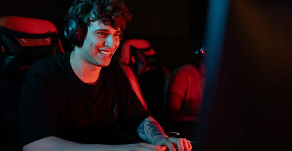 A young man wearing headphones and a black t-shirt is sitting in a dark room lit with red and blue light, looking at a computer screen and typing on a keyboard.