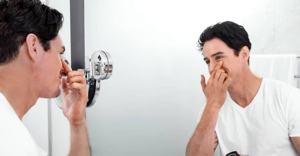 Man in white shirt in a bathroom applying face cream while looking in the mirror.