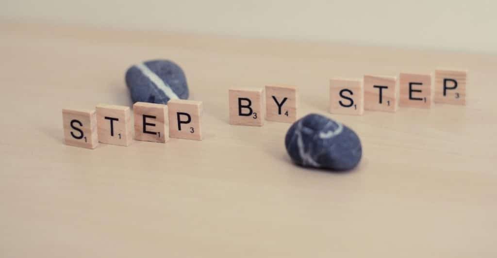 Wooden blocks spelling "STEP BY STEP" are arranged on a light surface, interspersed with small, smooth stones.