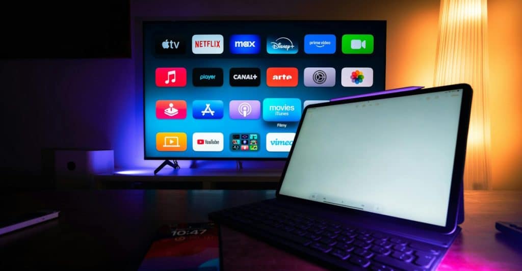 A laptop and a smartphone are on a table in front of a TV screen displaying various streaming service icons.