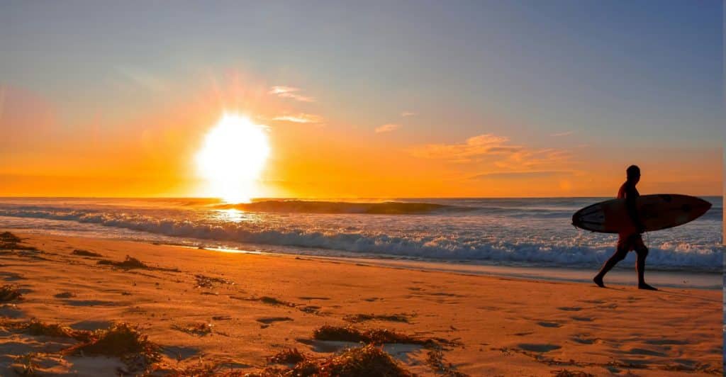 A silhouette of a person carrying a surfboard walking on a beach at sunset.