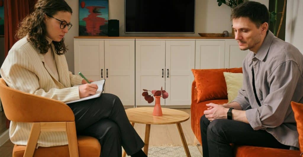  A therapist with curly hair taking notes while sitting across from a male client in a casual office setting.