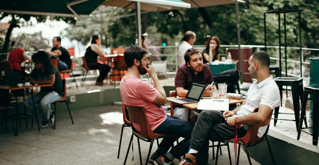 Three men sitting around a table outdoors at a cafe, talking and using a laptop.