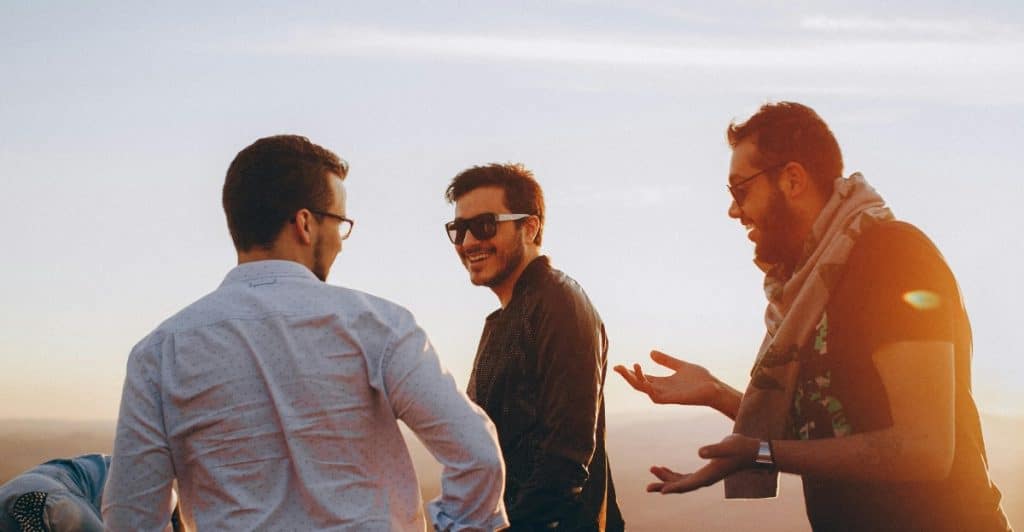 Three men stand outdoors talking and gesturing, with the sun setting behind them casting a warm glow.
