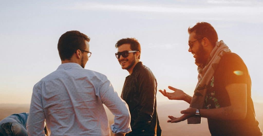 Three men standing outdoors on a hill during sunset, talking and gesturing with their hands, with a warm glow from the setting sun behind them.