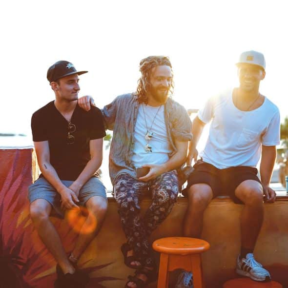Three men are sitting on a ledge outside, engaged in conversation, with a painted wall and stools visible.