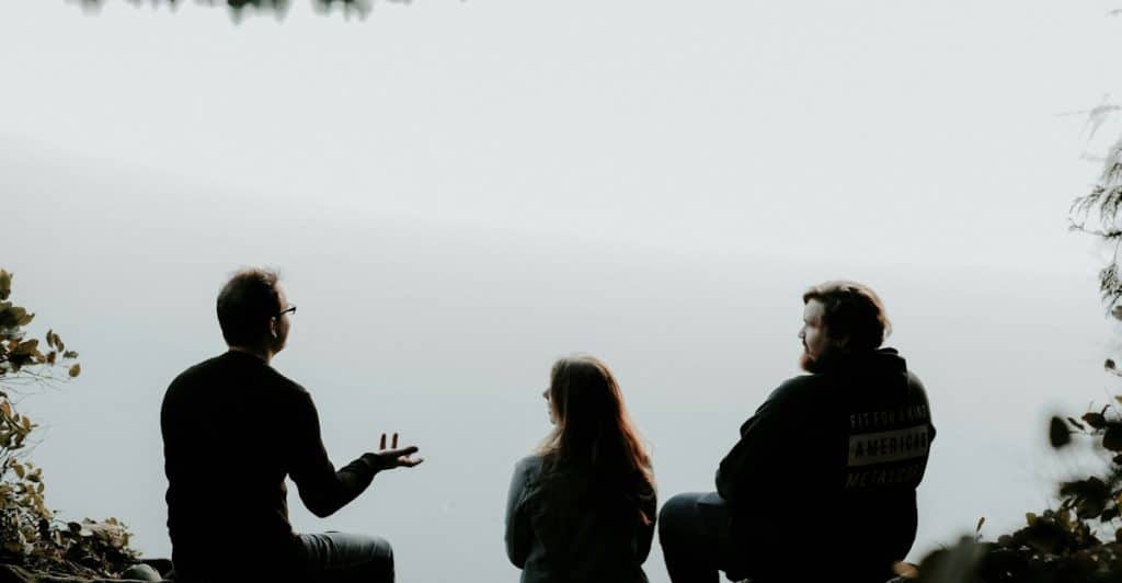 Three people sitting outdoors by a body of water, having a discussion.