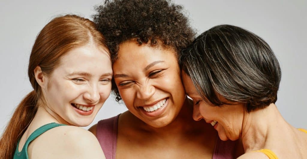 Three women with different hair colors are smiling and embracing