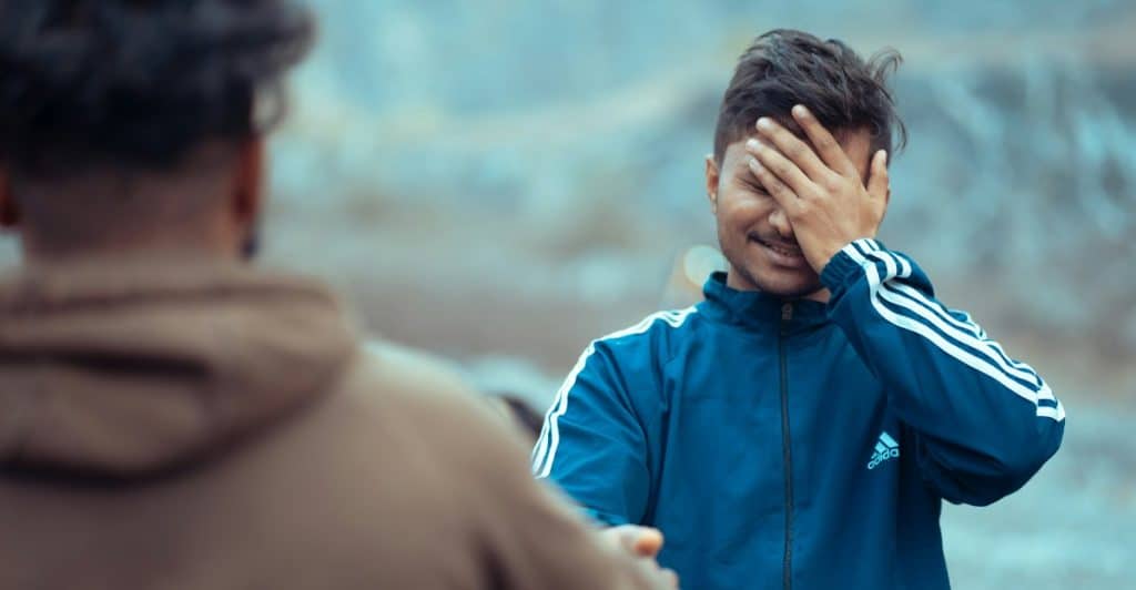 Two people outdoors, one reaching out a hand towards the other who is covering his face and smiling.