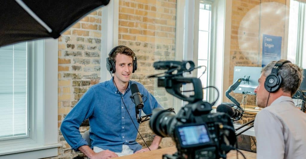 Two men sitting at a table with microphones and recording equipment, with a camera on a tripod in the foreground, filming the man in the blue shirt. The background shows an office or studio setting with brick walls and windows.
