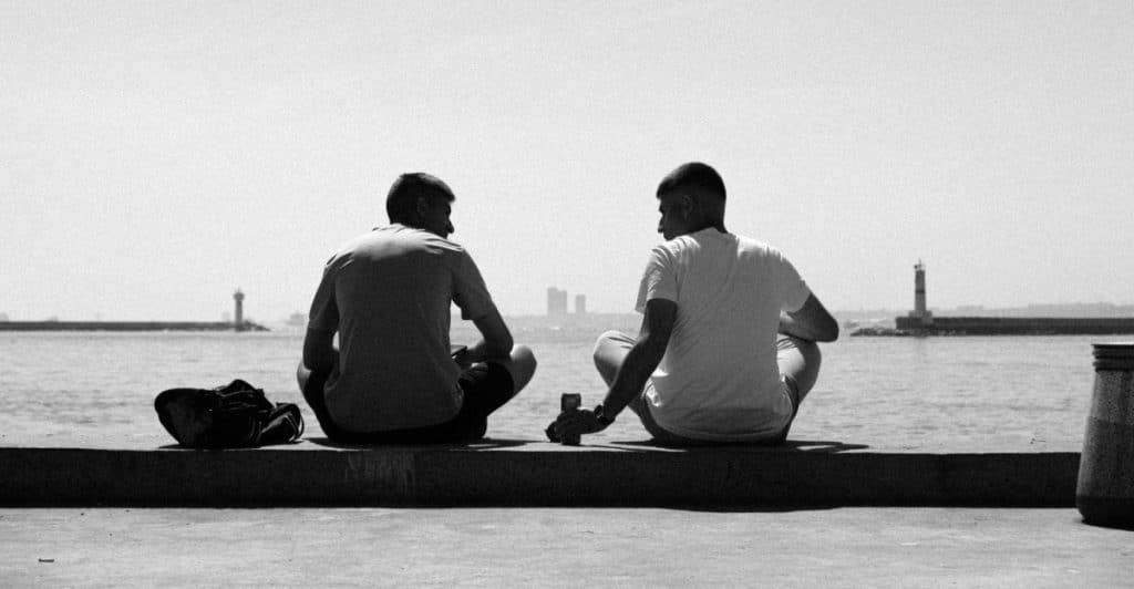 Two men sitting side-by-side on a ledge by the water, silhouetted against a bright sky.
