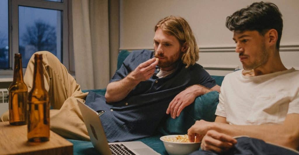 Two men sitting on a couch in a living room, watching a laptop and eating snacks from bowls.