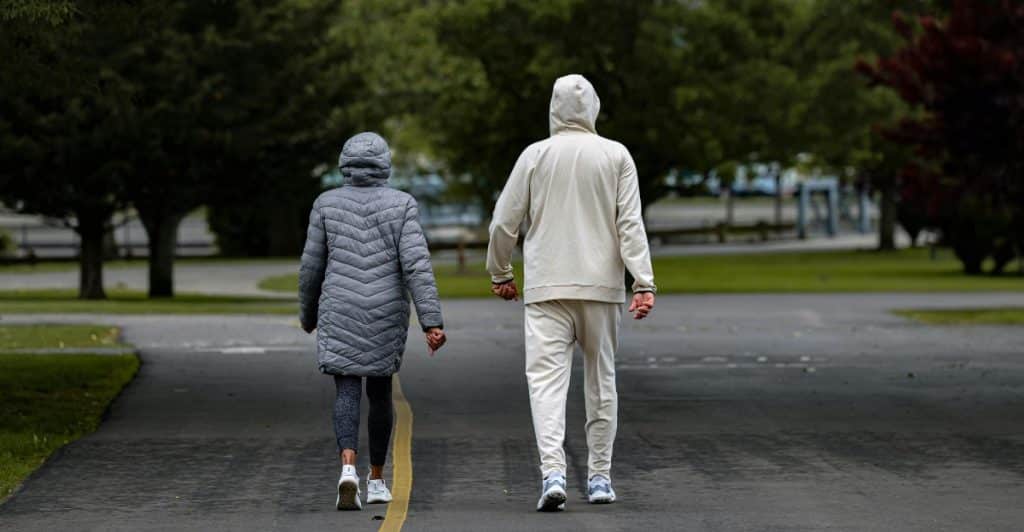 Two people wearing jackets are walking away from the viewer on a paved path in a green, outdoor setting.