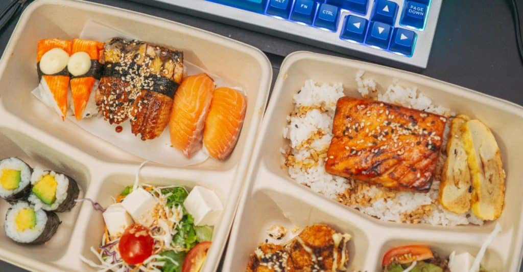 Two take-out containers filled with sushi, salad, rice, and other food items, placed on a surface next to a computer keyboard.