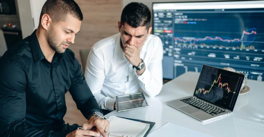 Two men reviewing financial charts on a laptop and a large screen in an office setting.