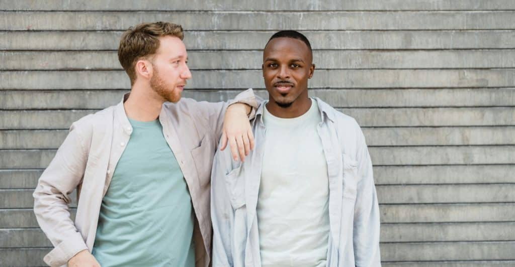 Two men are standing together outdoors against a textured gray wall, wearing light-colored layered outfits.