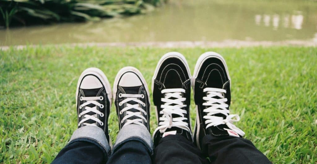  A close-up of a couple wearing two pairs of black and white sneakers sitting on a grassy surface.