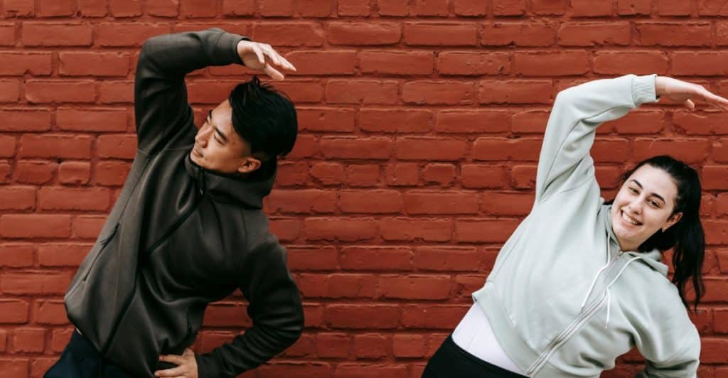 Two people in athletic wear stretching their arms in an outdoor setting with a brick wall behind them.