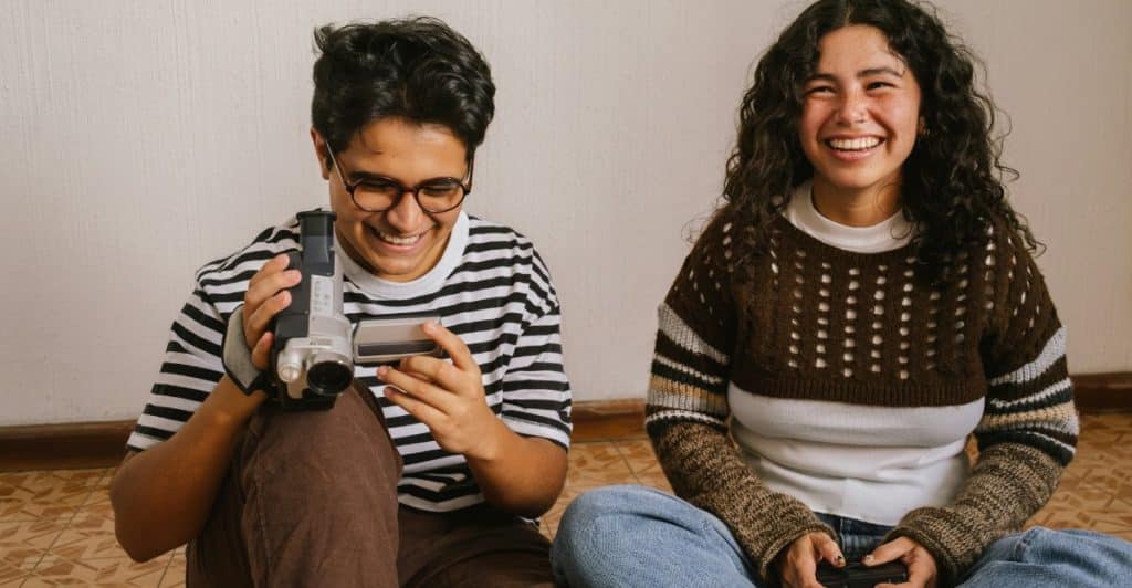 A young man with glasses holding a video camera and smiling with a young woman who is also smiling, sitting on the floor.