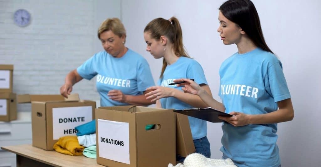 Three women wearing blue volunteer shirts sorting clothing items into cardboard donation boxes.